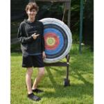 Teenager in a dark hoodie and black shorts points at a large circular archery target on a wooden stand outdoors.