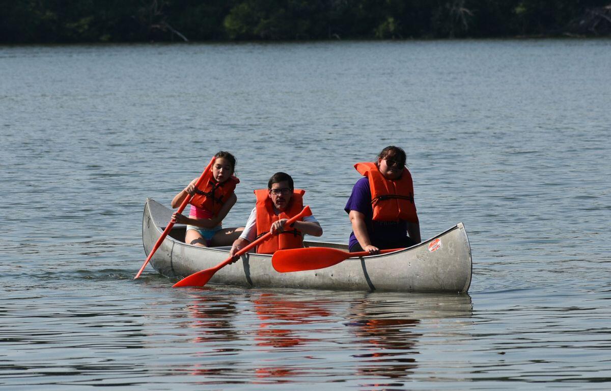 Three people in a canoe on calm water wearing bright orange life jackets, paddling with red oars.