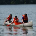 Three people in a canoe on calm water wearing bright orange life jackets, paddling with red oars.