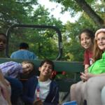 Group of diverse kids riding in the back of a green trailer outdoors, smiling at the camera.