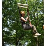 Young woman wearing a helmet and harness zip-lining through a forested area, smiling on the ride.