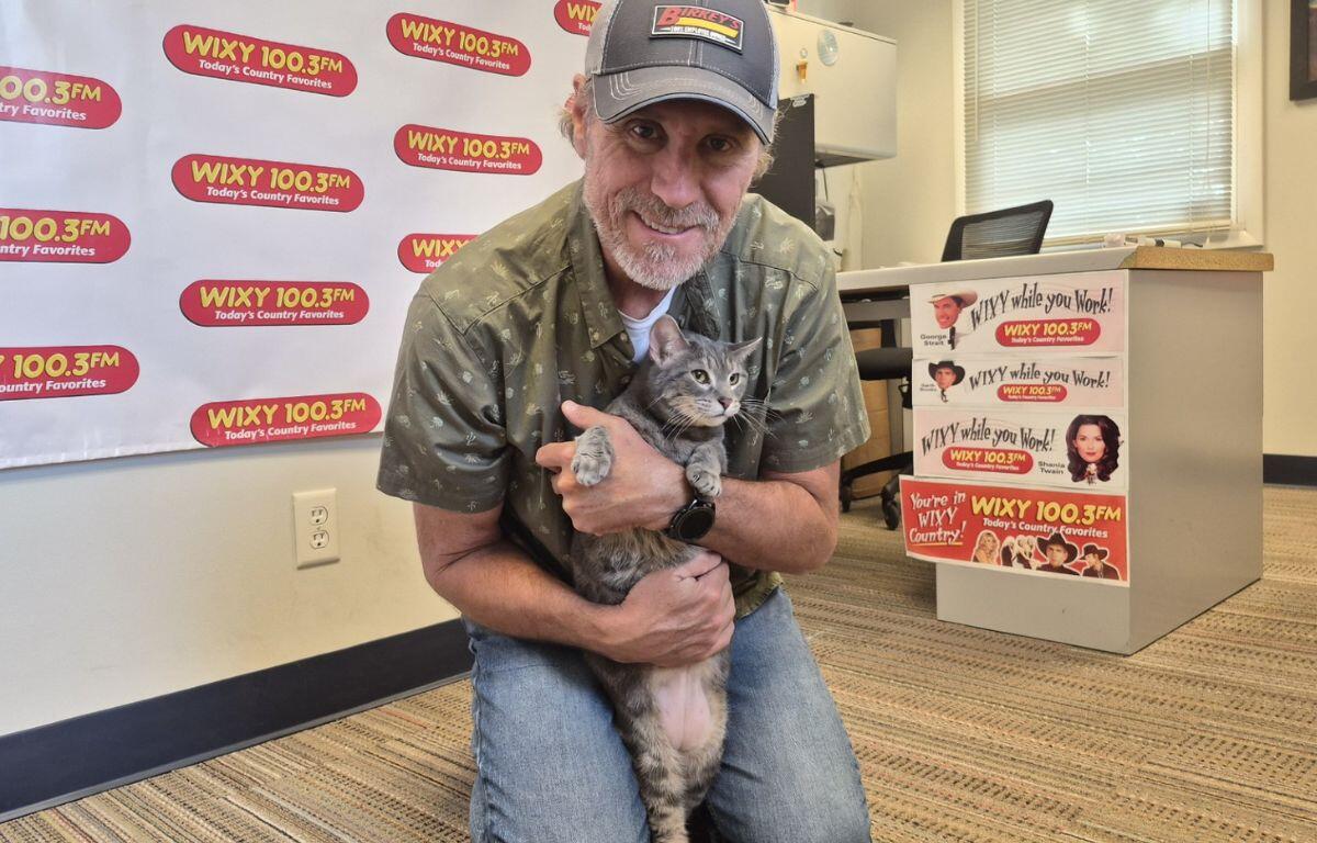 Man wearing a cap hugs a gray cat in a radio station studio with WIXY 100.3FM banners in the background