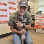 Man wearing a cap hugs a gray cat in a radio station studio with WIXY 100.3FM banners in the background