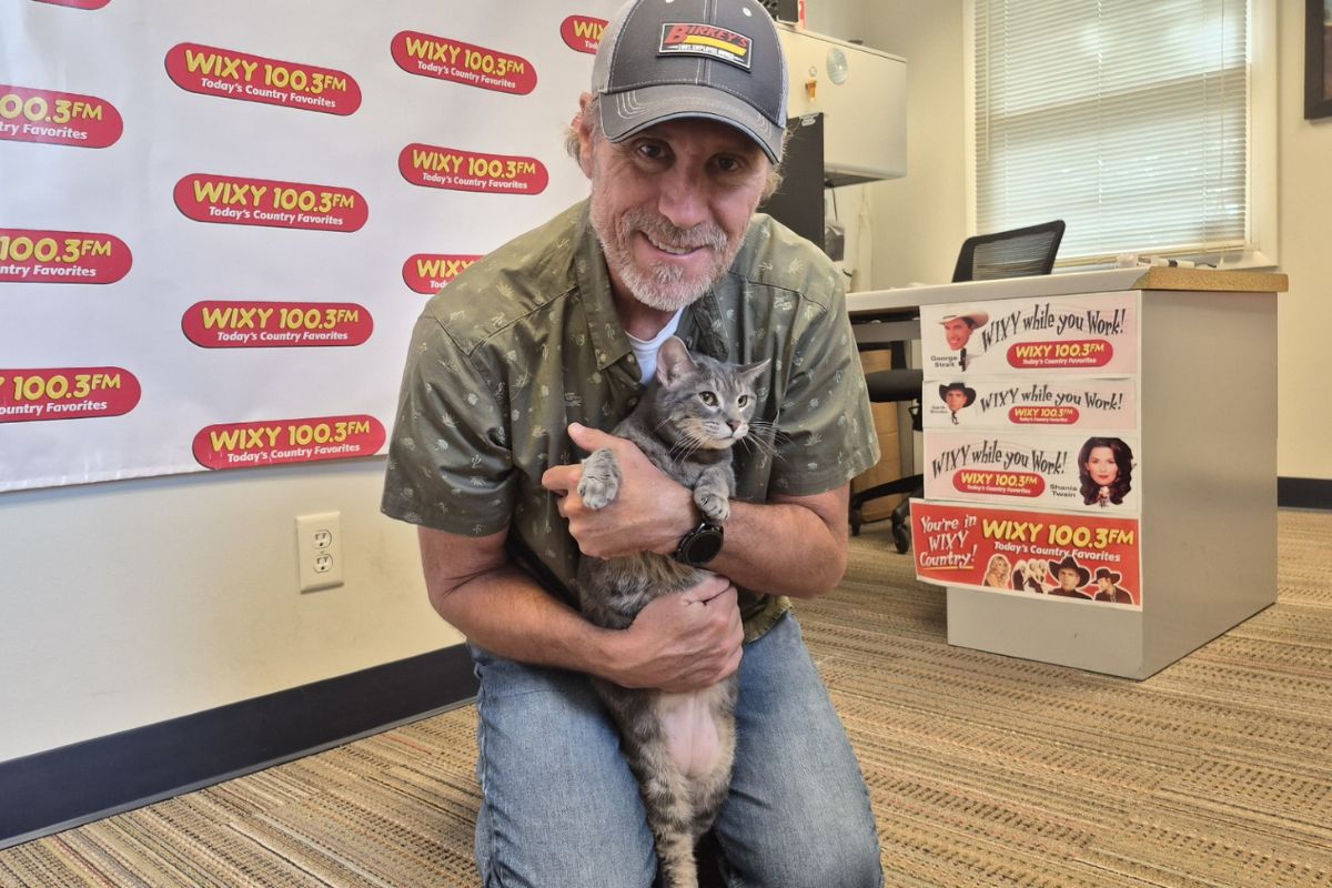 Man wearing a cap hugs a gray cat in a radio station studio with WIXY 100.3FM banners in the background