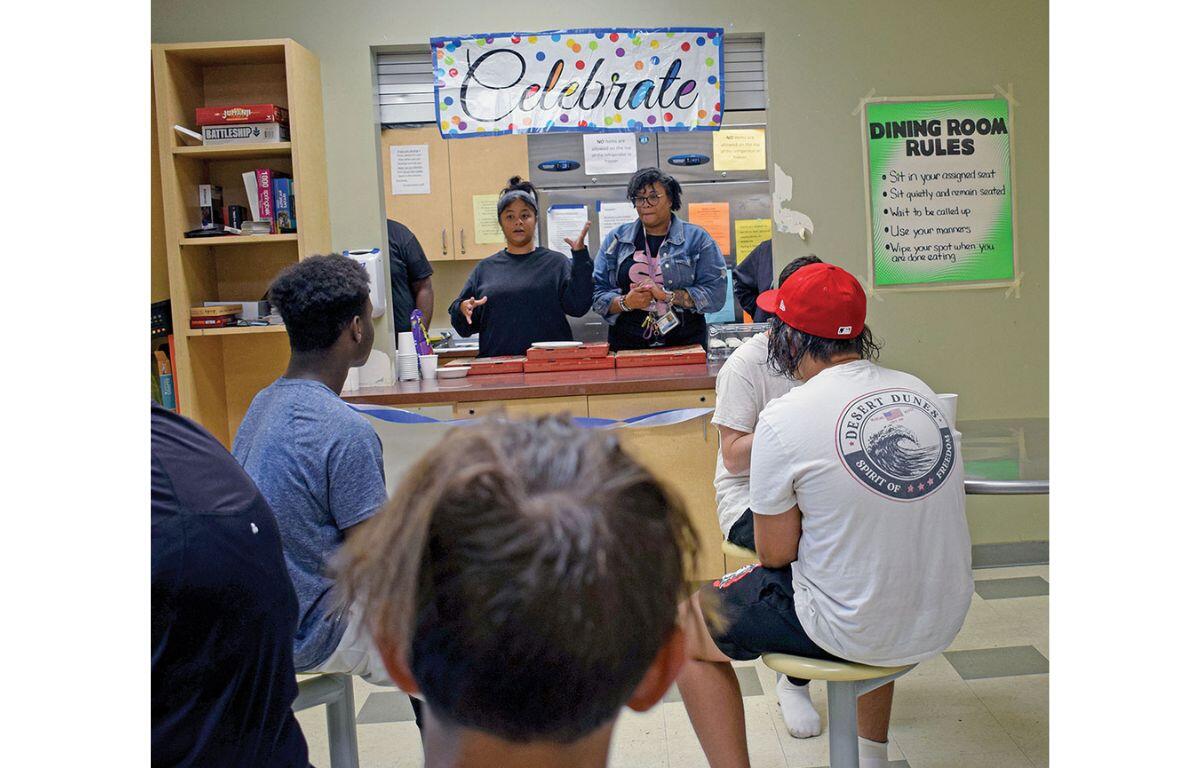 Students sit at a cafeteria counter as two staff members hand out pizza boxes under a colorful Celebrate banner above them.