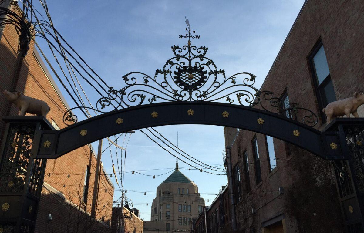 Ornate wrought-iron arch gate with scrollwork and animal sculptures, opening to a brick street with a domed building in the distance.