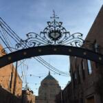 Ornate wrought-iron arch gate with scrollwork and animal sculptures, opening to a brick street with a domed building in the distance.