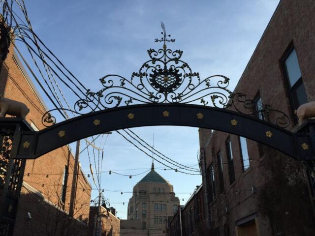 Ornate wrought-iron arch gate with scrollwork and animal sculptures, opening to a brick street with a domed building in the distance.