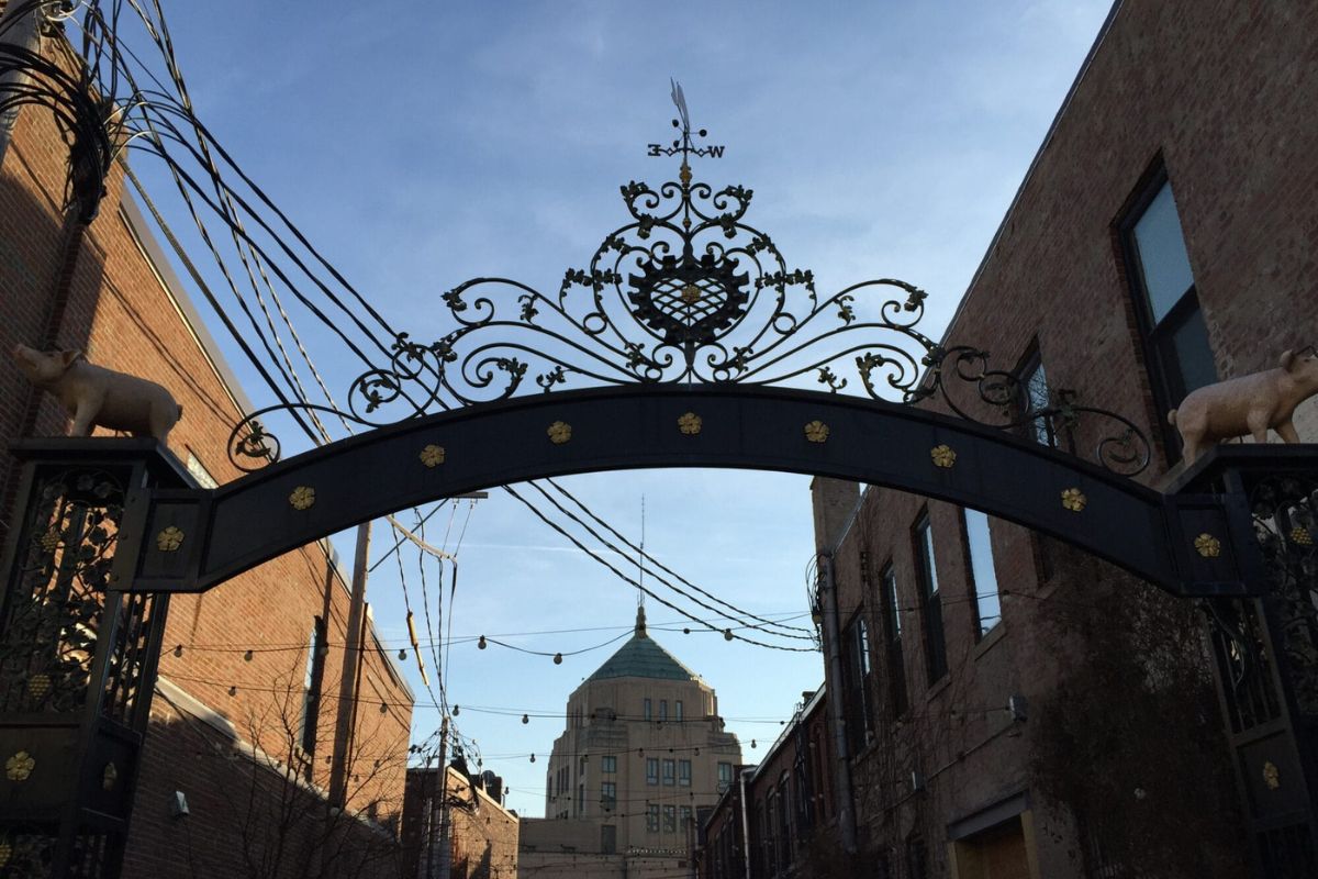 Ornate wrought-iron arch gate with scrollwork and animal sculptures, opening to a brick street with a domed building in the distance.