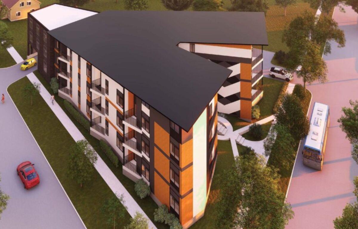 Aerial view of a modern multi-story apartment building with orange panels, balconies, and a dark slanted roof surrounded by trees and roads.