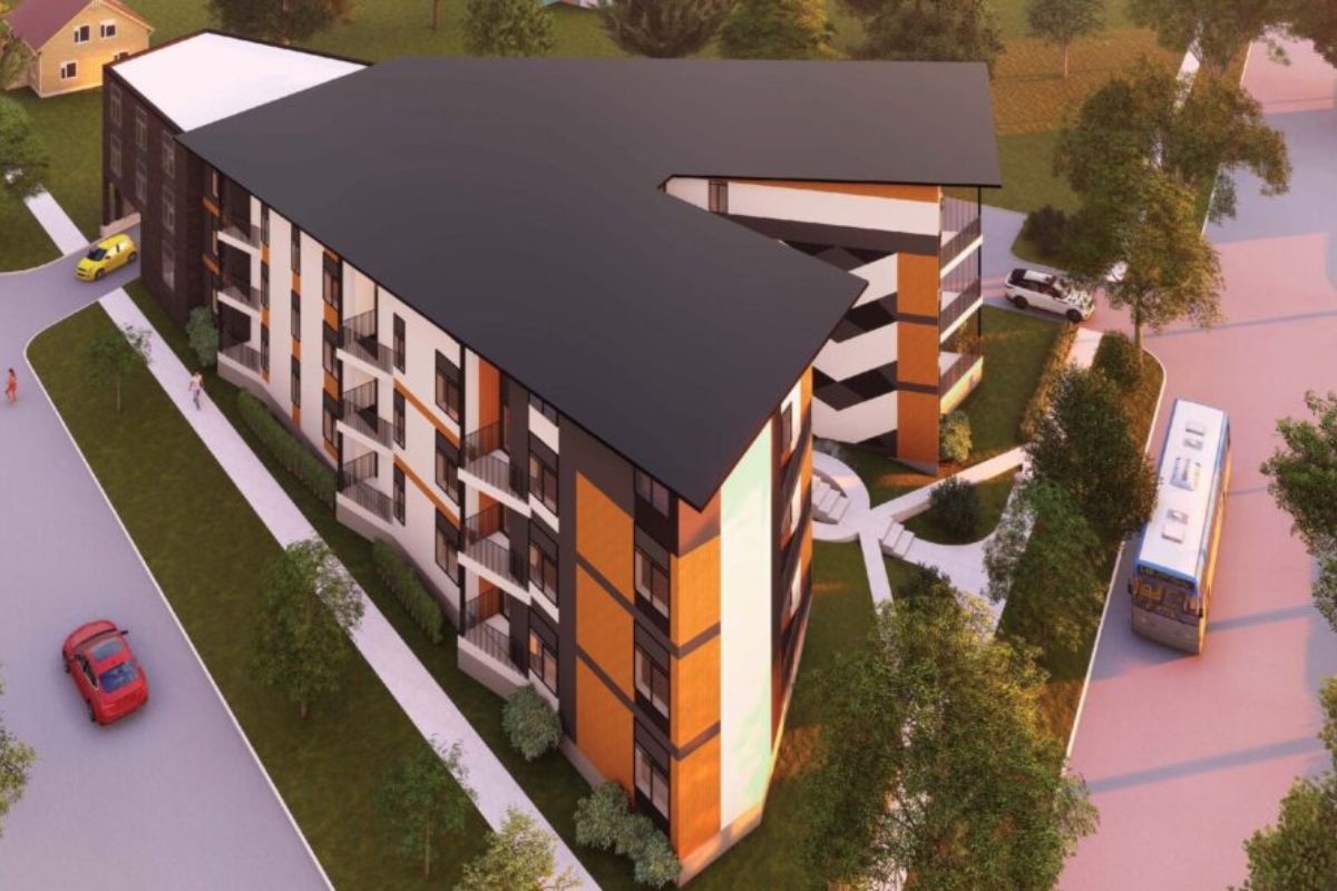 Aerial view of a modern multi-story apartment building with orange panels, balconies, and a dark slanted roof surrounded by trees and roads.