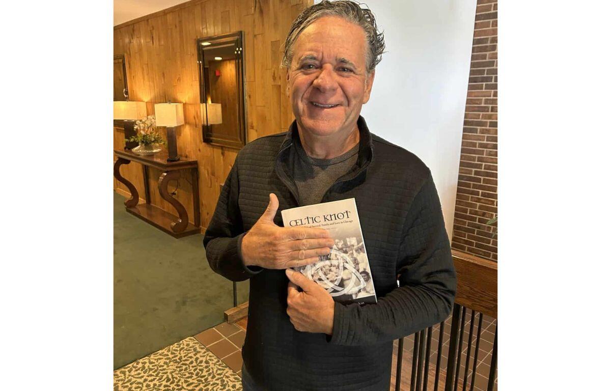 Smiling man in a dark quilted sweater holds a book titled Celtic Knot in a warmly lit lobby backdrop.