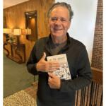 Smiling man in a dark quilted sweater holds a book titled Celtic Knot in a warmly lit lobby backdrop.