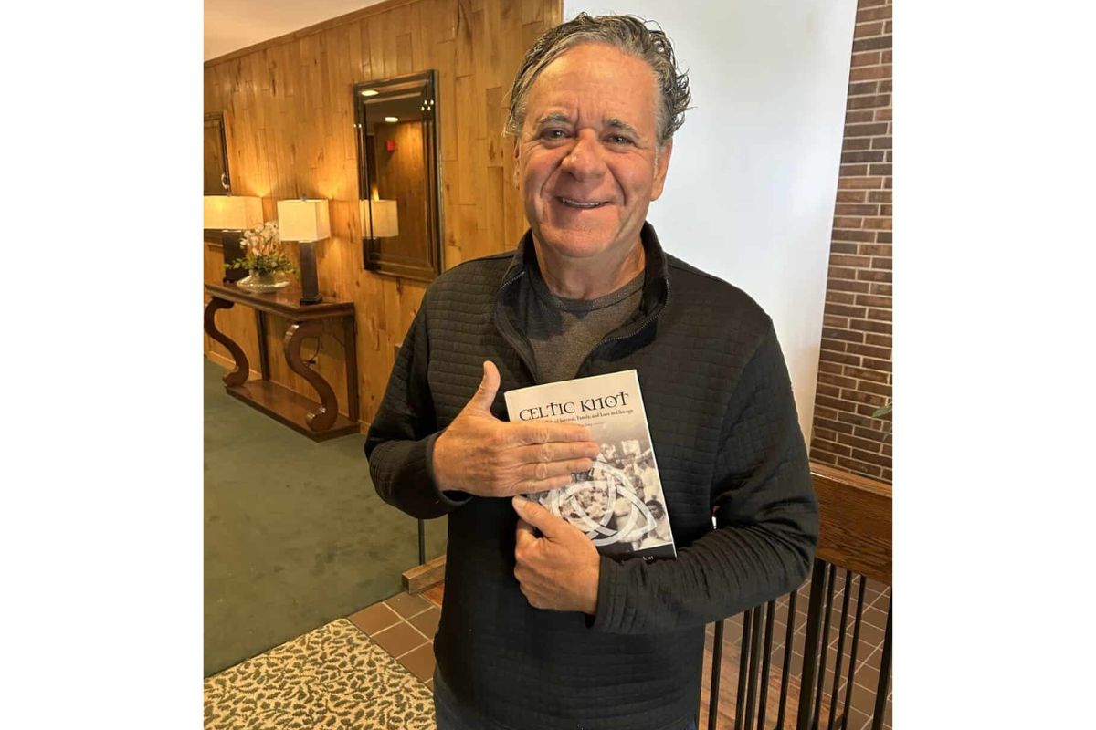 Smiling man in a dark quilted sweater holds a book titled Celtic Knot in a warmly lit lobby backdrop.