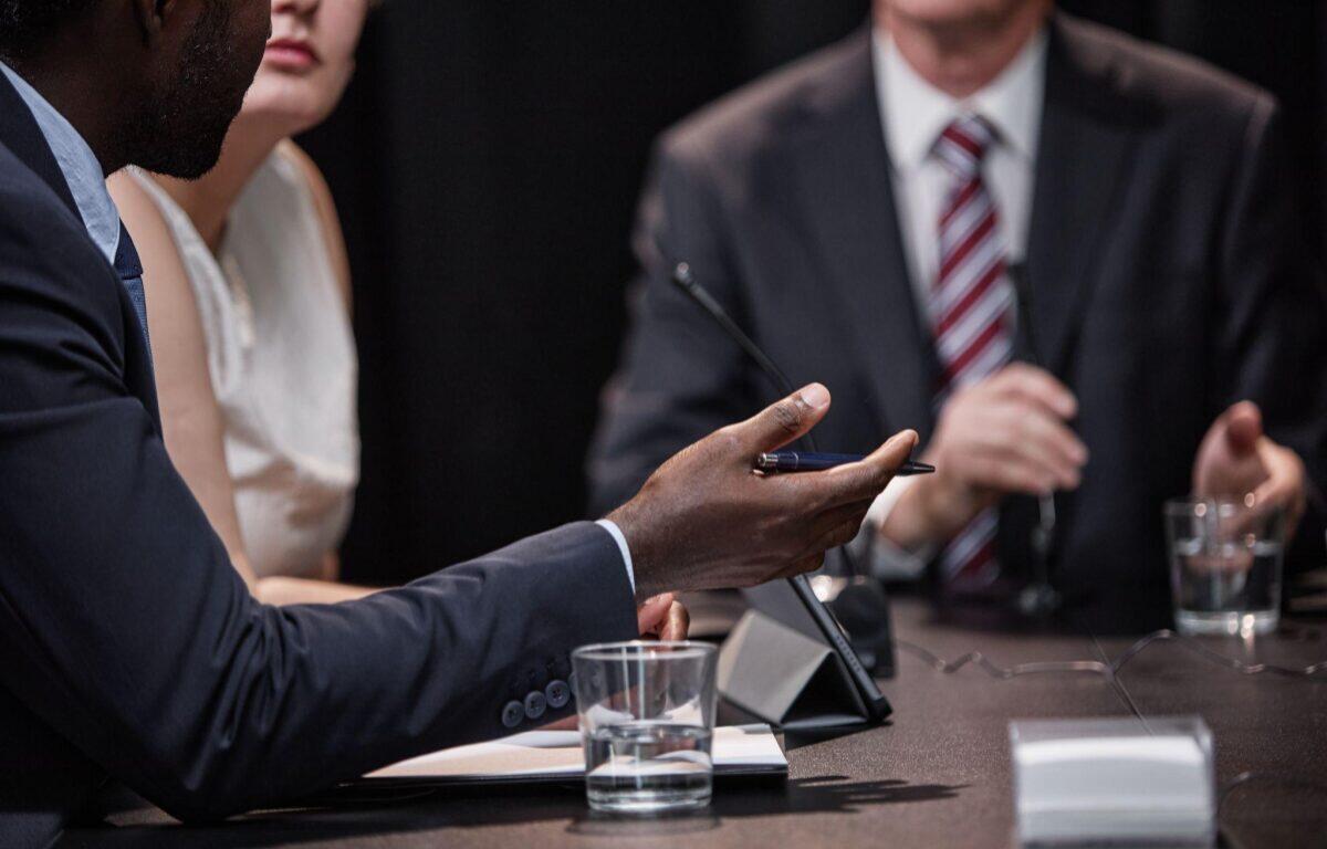 Close-up of a man in a navy suit gesturing with a pen during a business meeting at a conference table.