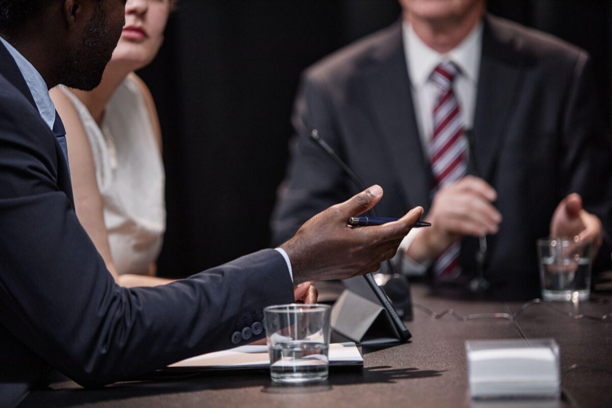 Close-up of a man in a navy suit gesturing with a pen during a business meeting at a conference table.