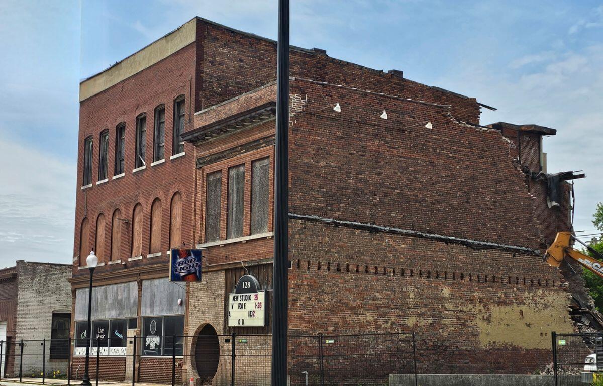 Brick building with boarded storefronts and a partially demolished upper level against a blue sky, construction in progress on the right side.
