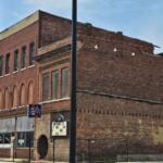 Brick building with boarded storefronts and a partially demolished upper level against a blue sky, construction in progress on the right side.