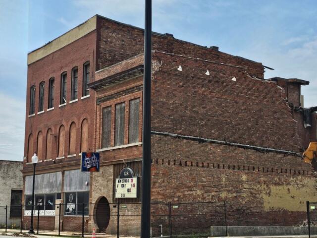 Brick building with boarded storefronts and a partially demolished upper level against a blue sky, construction in progress on the right side.