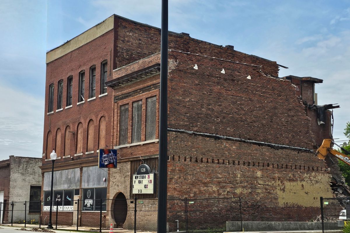 Brick building with boarded storefronts and a partially demolished upper level against a blue sky, construction in progress on the right side.