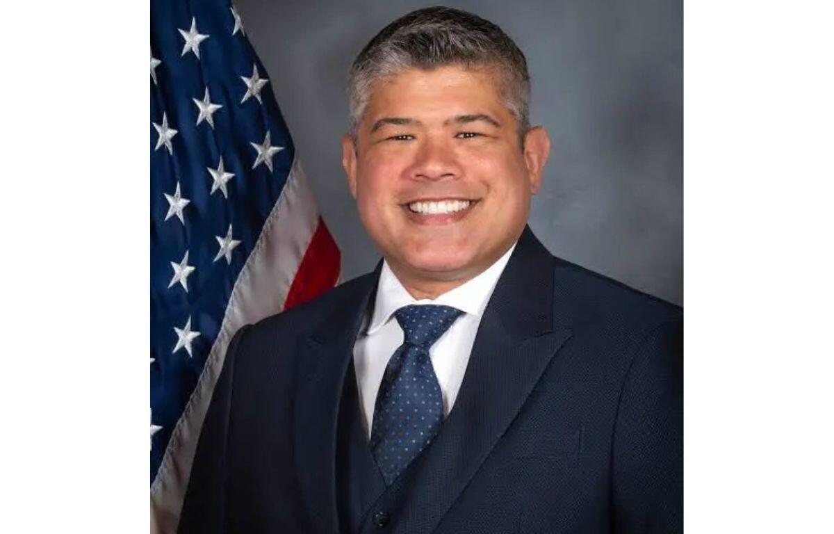 Smiling man in a dark suit and blue tie, posing in front of the American flag.