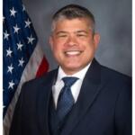 Smiling man in a dark suit and blue tie, posing in front of the American flag.