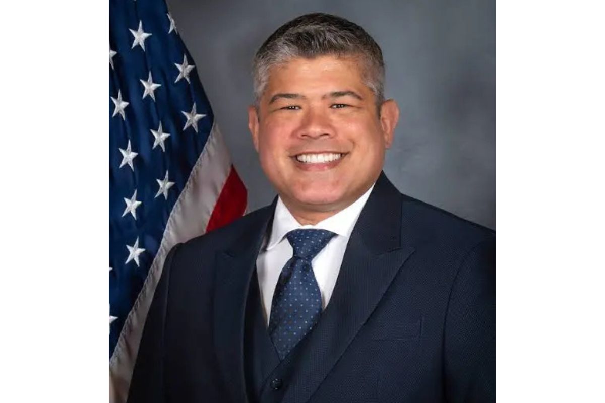 Smiling man in a dark suit and blue tie, posing in front of the American flag.