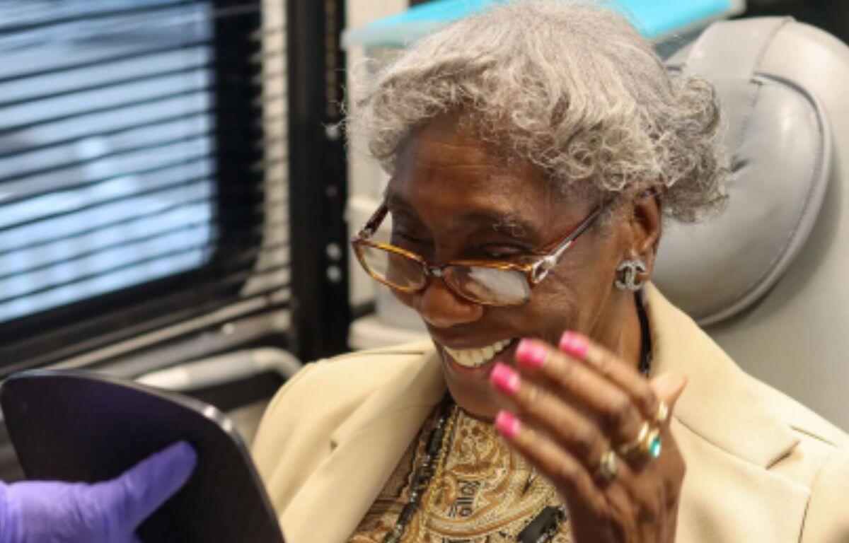 Elderly woman with gray hair and glasses smiling while looking at a tablet handed by a person in a purple glove in a clinical setting