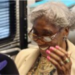 Elderly woman with gray hair and glasses smiling while looking at a tablet handed by a person in a purple glove in a clinical setting
