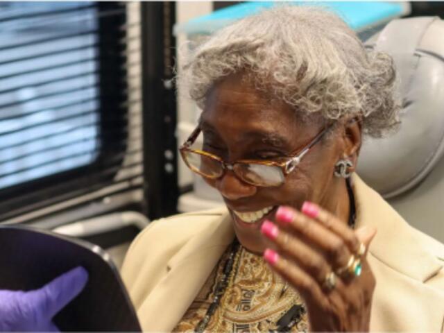 Elderly woman with gray hair and glasses smiling while looking at a tablet handed by a person in a purple glove in a clinical setting