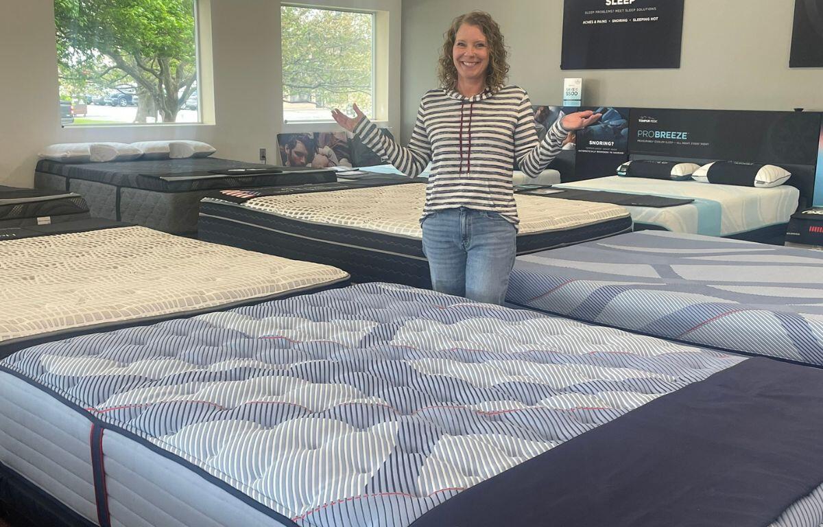 Smiling woman stands in a mattress showroom with arms outstretched among rows of patterned beds.