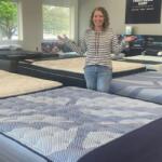 Smiling woman stands in a mattress showroom with arms outstretched among rows of patterned beds.