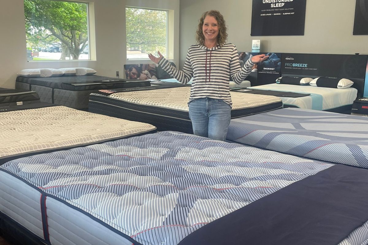 Smiling woman stands in a mattress showroom with arms outstretched among rows of patterned beds.