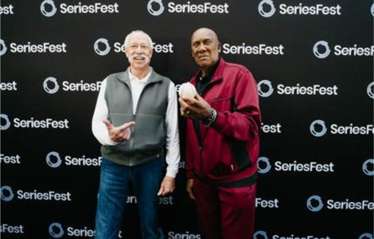 Two smiling men pose together in front of a SeriesFest backdrop; the man on the right holds a white helmet.