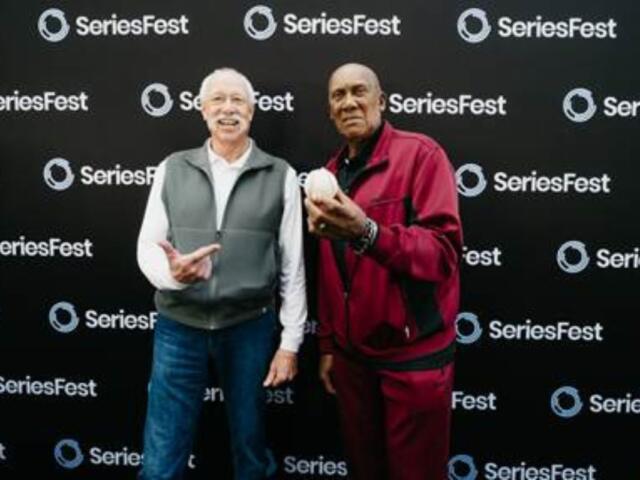 Two smiling men pose together in front of a SeriesFest backdrop; the man on the right holds a white helmet.