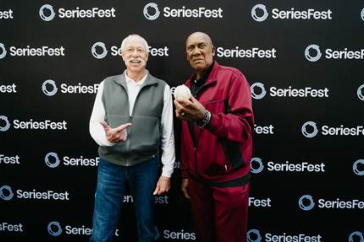 Two smiling men pose together in front of a SeriesFest backdrop; the man on the right holds a white helmet.