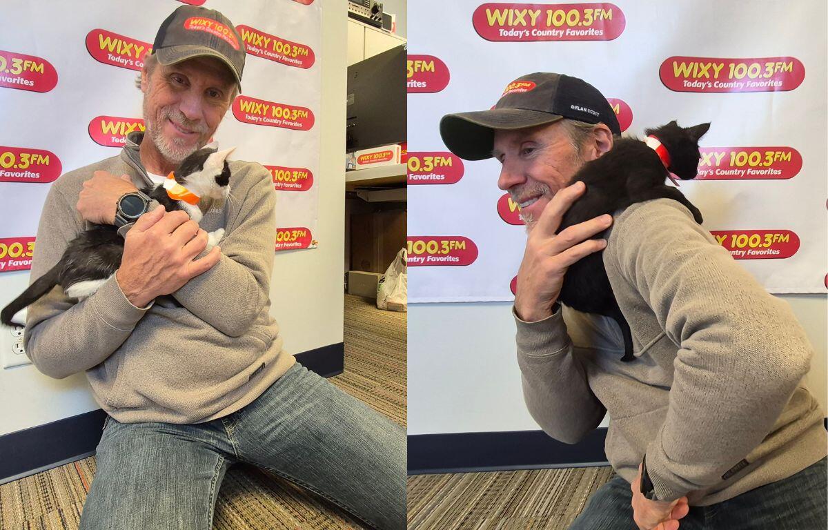 Man in a gray hoodie and cap sitting on the floor, gently holding a black-and-white cat with an orange bow, in front of a WXKY 100.3FM banner.