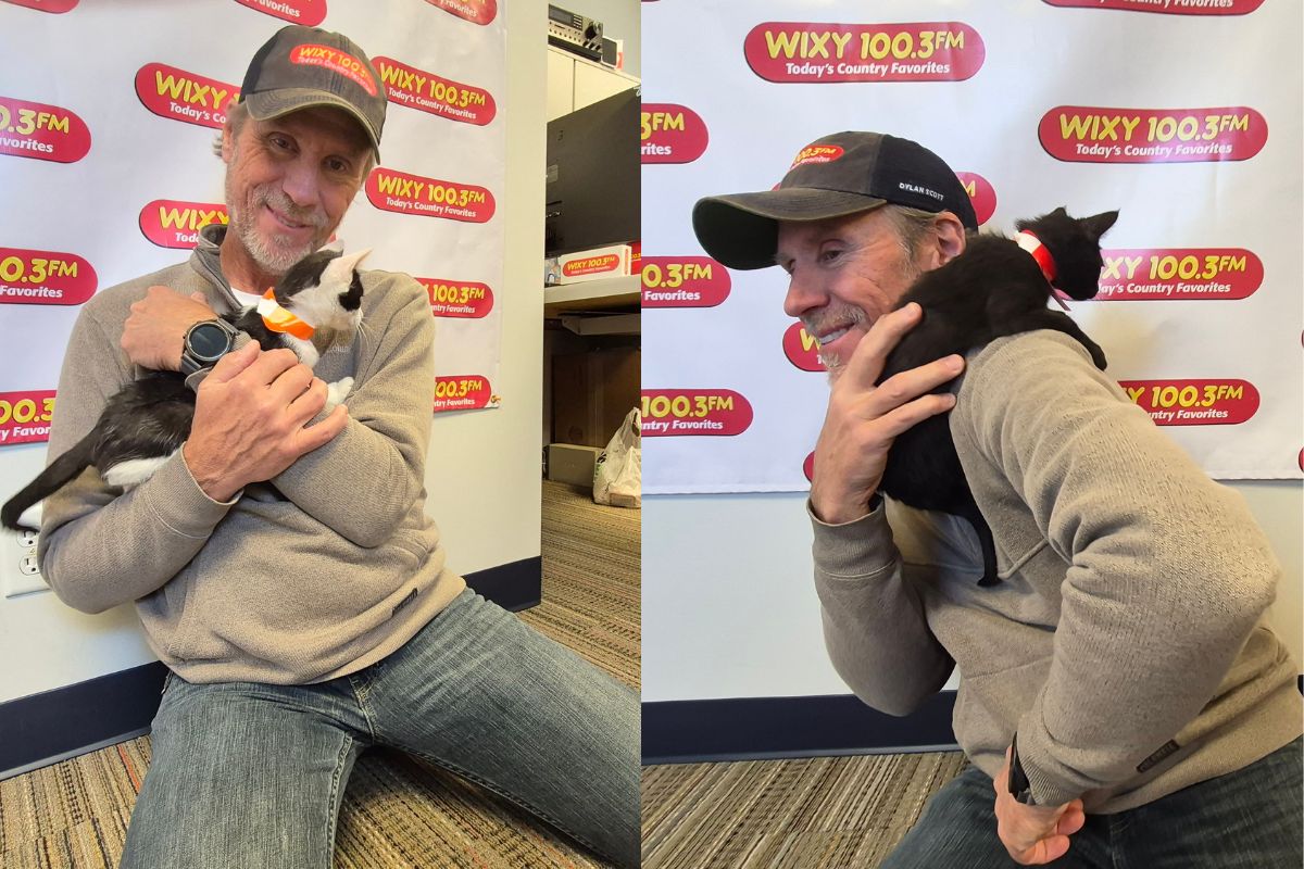 Man in a gray hoodie and cap sitting on the floor, gently holding a black-and-white cat with an orange bow, in front of a WXKY 100.3FM banner.