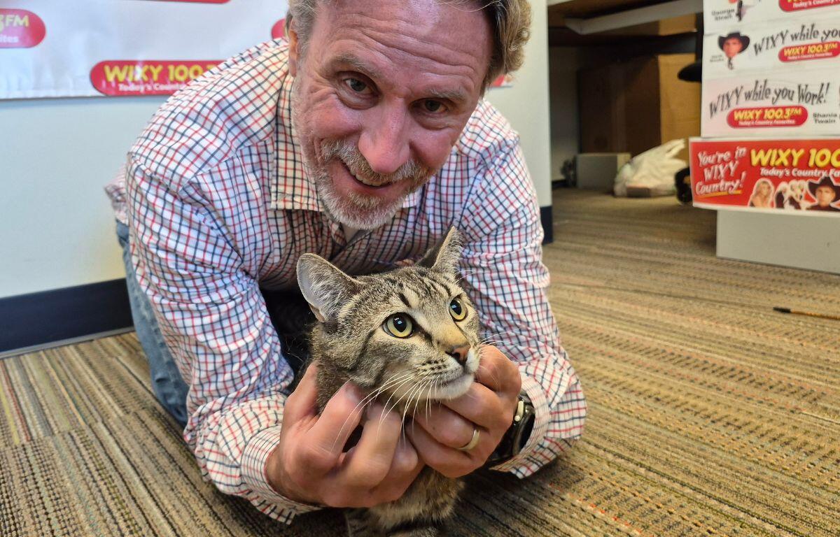 Man in a plaid shirt smiles while holding a tabby cat on a striped office carpet with WIXY banners in the background, likely a radio station setting.