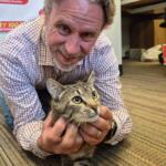 Man in a plaid shirt smiles while holding a tabby cat on a striped office carpet with WIXY banners in the background, likely a radio station setting.