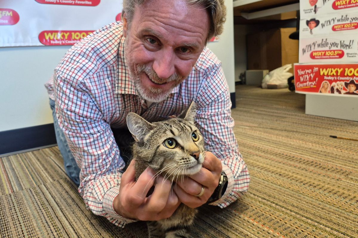 Man in a plaid shirt smiles while holding a tabby cat on a striped office carpet with WIXY banners in the background, likely a radio station setting.
