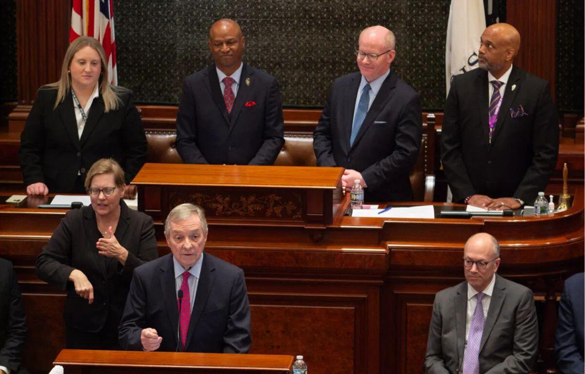 Man in a dark suit speaks from a wooden podium in a formal chamber, with colleagues standing and seated behind him.