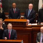 Man in a dark suit speaks from a wooden podium in a formal chamber, with colleagues standing and seated behind him.