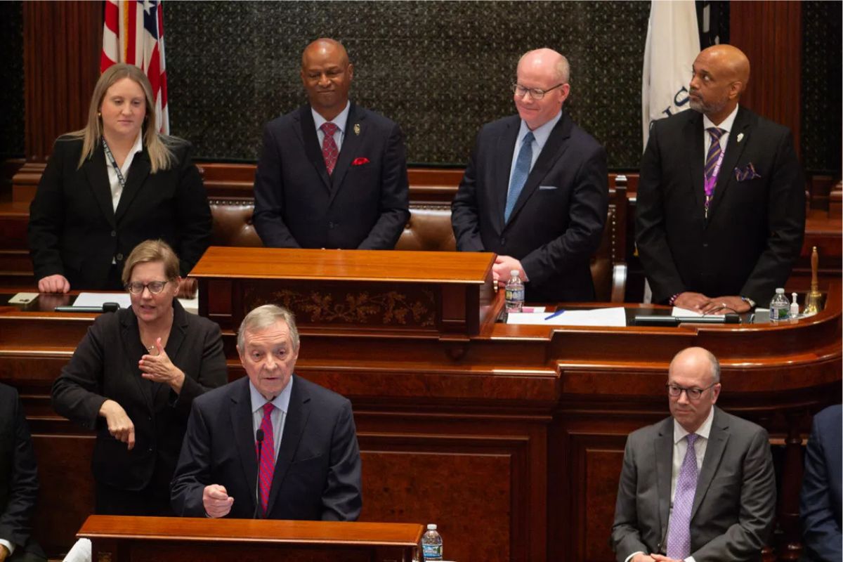 Man in a dark suit speaks from a wooden podium in a formal chamber, with colleagues standing and seated behind him.