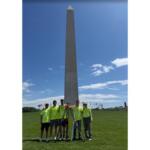 Group of six people in neon shirts posing with surveying equipment in front of the Washington Monument on the National Mall.