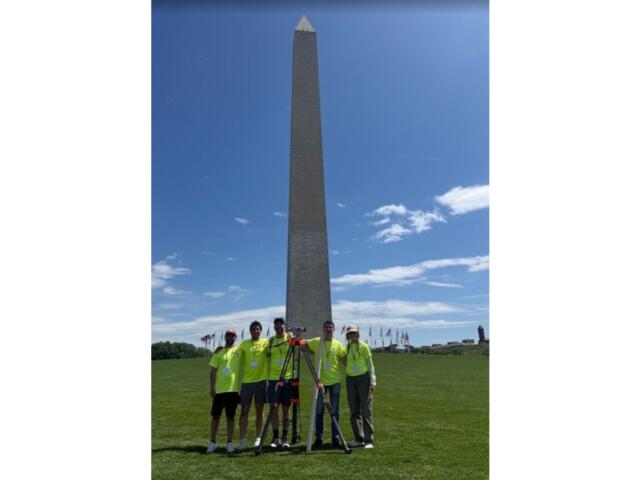 Group of six people in neon shirts posing with surveying equipment in front of the Washington Monument on the National Mall.