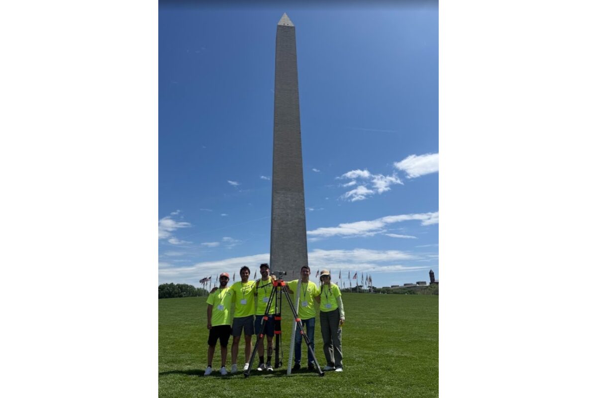 Group of six people in neon shirts posing with surveying equipment in front of the Washington Monument on the National Mall.