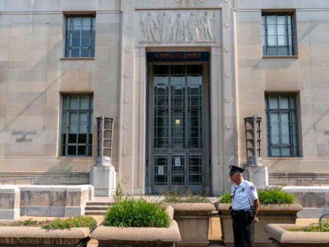 Front entrance of a government building with a security guard standing near planters and steps.