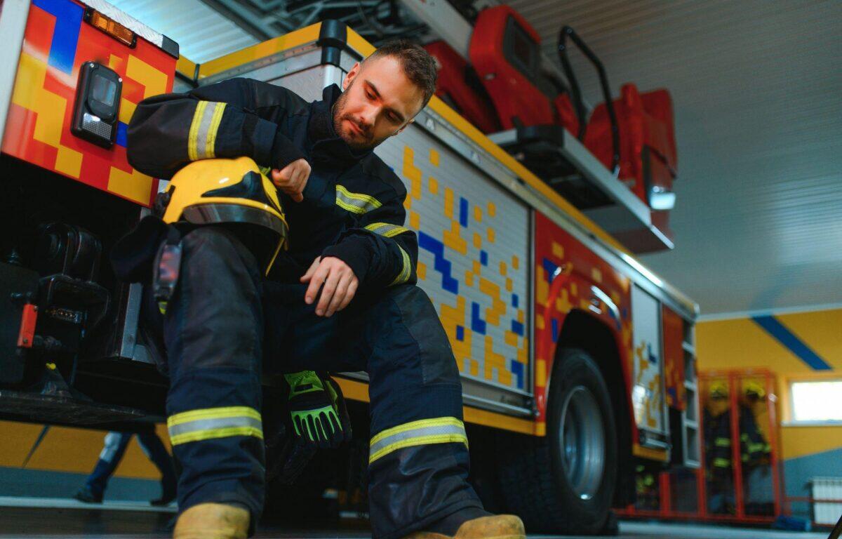 Firefighter in full gear sitting by a colorful fire truck, putting on or inspecting a helmet in a station bay.
