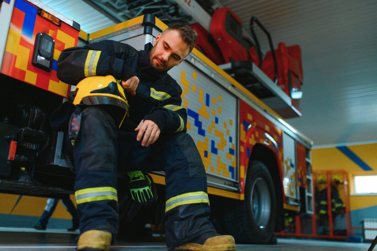 Firefighter in full gear sitting by a colorful fire truck, putting on or inspecting a helmet in a station bay.